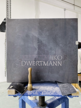 A gravestone made of dark Irish limestone in the workshop. The rough surface of the stone has been slightly cleaned, and the name is engraved on it. A wooden mallet is resting on the stone.