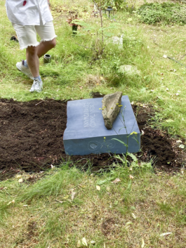 The flat gravestone made of dark Irish limestone is installed on a lawn grave at Friedrichswerder Cemetery. A small stone sculpture and flowers are placed on the grave, and a person is standing in the background.